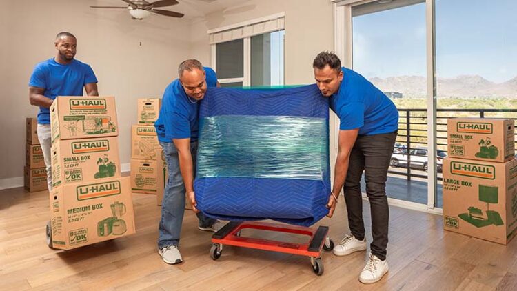 Two Moving Helpers place a piece of furniture wrapped in furniture blankets and stretch wrap onto a furniture dolly. Another Moving Help Service Provider pushes a dolly with three moving boxes on the dolly next to them